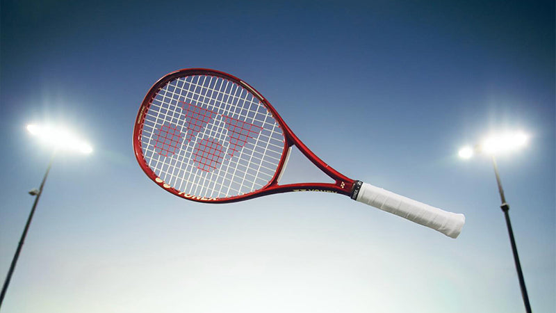 Red Yonex VCORE tennis racquet floating in the  air against a clear sky background with bright court lights turned on.