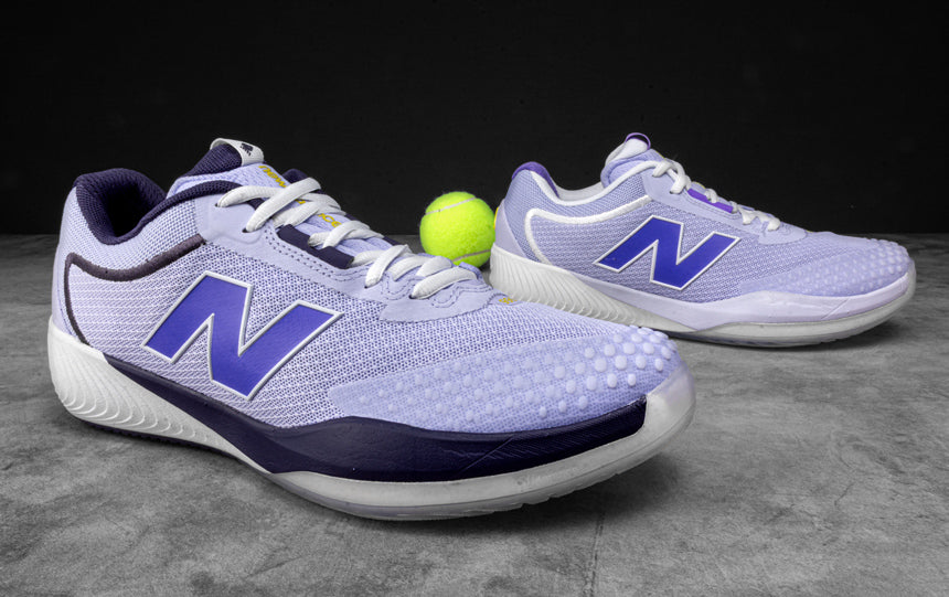Pair of purple and navy New Balance tennis shoes with a tennis ball on a gray surface.