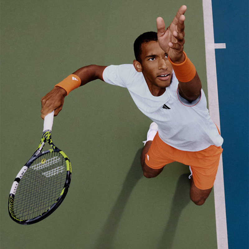 A person about to serve a tennis ball wearing white and orange adidas tennis clothing and shoes on a blue court