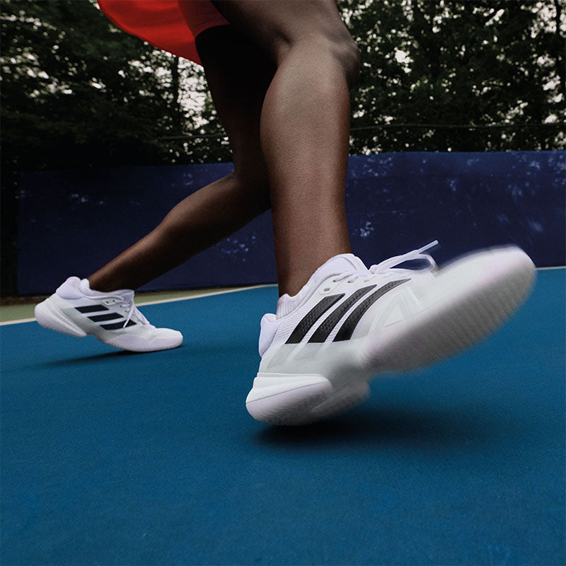 Closeup of a person playing tennis in white and black adidas Barricade tennis shoes on a blue court.