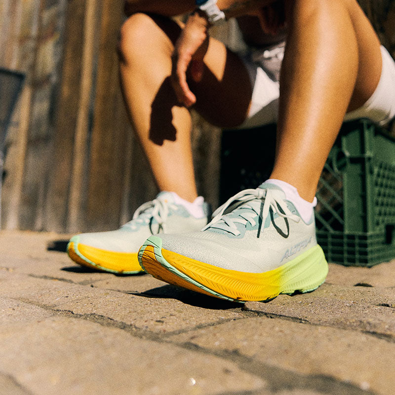 A person sitting on a green plastic crate wearing gray Altra Experience Flow 3 running shoes with orange and green-yellow accents on a brick sidewalk