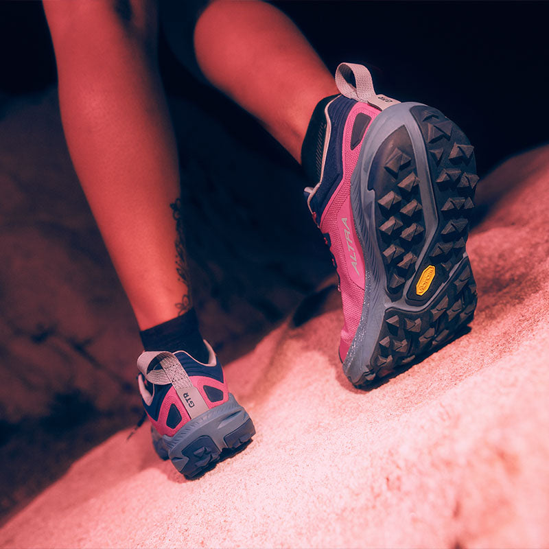 A person wearing pink Altra Experience Wild 3+ trail running shoes with purple accents on a rocky surface