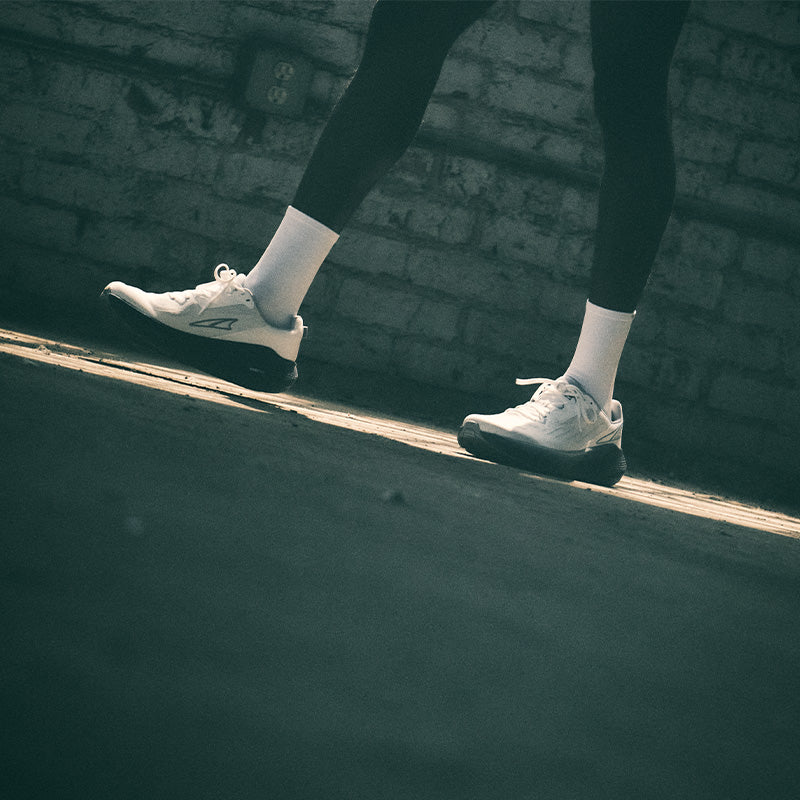 Person wearing white Altra FWD Via running shoes and dark leggings walking on a sidewalk with a brick wall in the background.