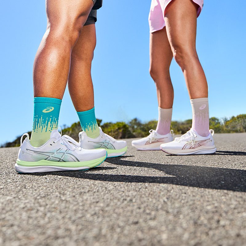 Two runners wearing ASICS GEL-Kayano 33 running shoes on a paved road with green bushes and a clear sky in the background