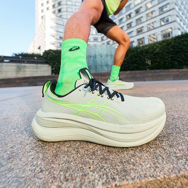 Person wearing a light colored ASICS GEL-Nimbus 28 men's running shoe with green accents on a city sidewalk.