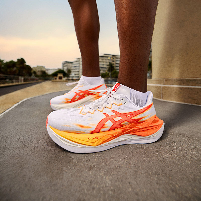Close-up of a runner wearing white ASICS Superblast 3 running shoes with bright orange and yellow accents on a concrete surface with trees and buildings in the background.