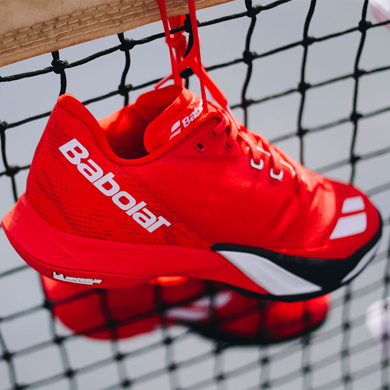 close up of a pair of red Babolat Jet Mach 4 tennis shoes hanging over a net  on a gray blue court.