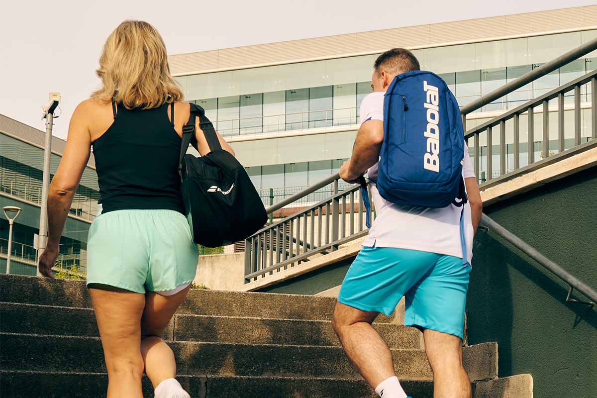 Two people walking up concrete stairs outdoors, one with a black Babolat Court S Padel Bag and the other with a blue Babolat Court Lite Padel Backpack.