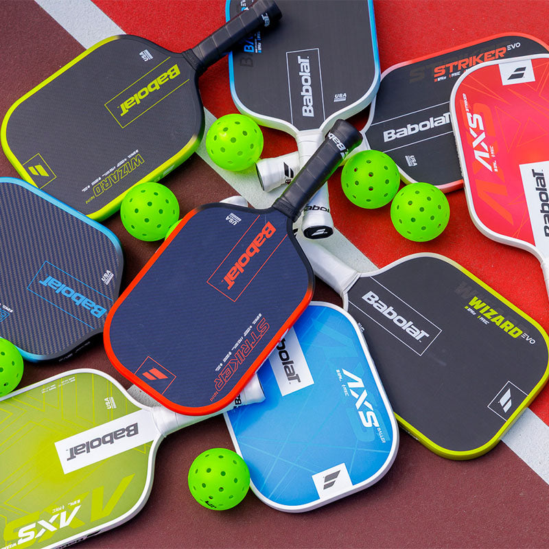 Various Babolat pickleball paddles scattered on a red court surface with some pickleballs