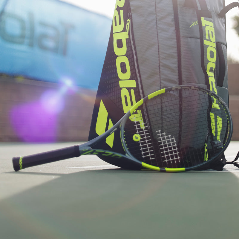 A dark blue-green Babolat Pure Aero 2026 tennis racquet with neon yellow-green accents leaning against a matching racquet bag on a green tennis court with blurred background.