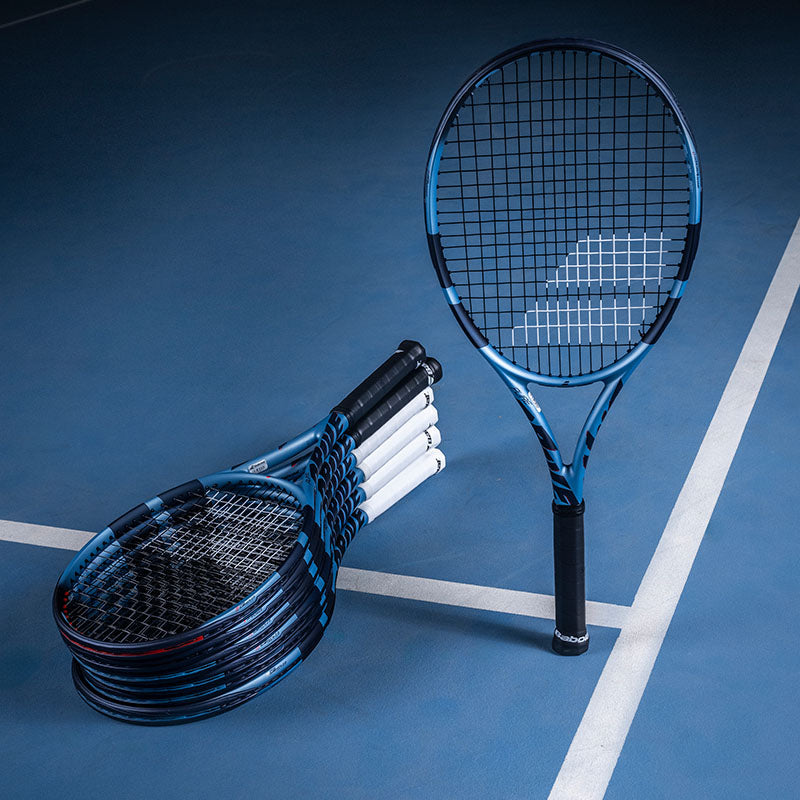 A set of Babolat Pure Drive tennis racquets in blue and black, with one standing upright and several stacked on a blue tennis court surface with white lines.