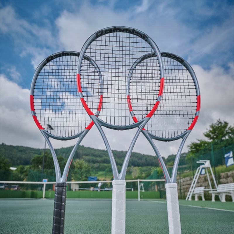 Three Babolat Pure Strike Carbon Grey tennis racquets stacked on a tennis court with a cloudy sky in the background