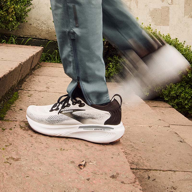 A person wearing gray pants and a white and black pair of Brooks Adrenaline GTS 25 running shoes walking up a stone stairway outdoors, with motion blur on the hind leg.