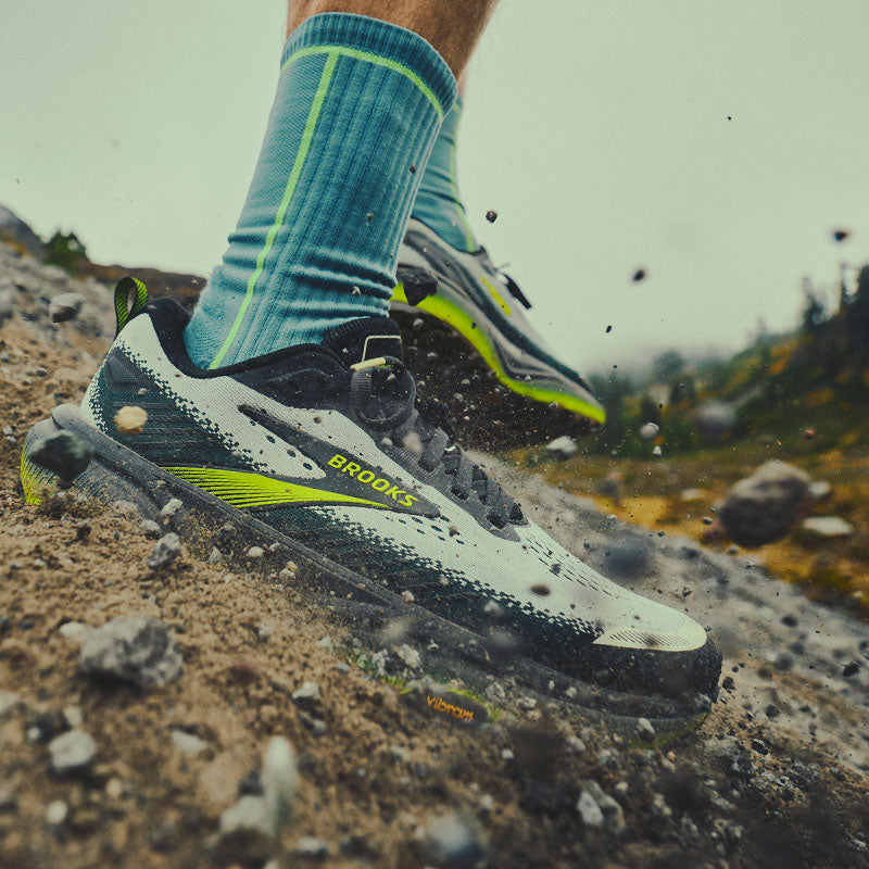 A close-up action shot of a person wearing Brooks Cascadia 20 trail running shoes and teal socks, kicking up dirt and rocks on an outdoor trail.