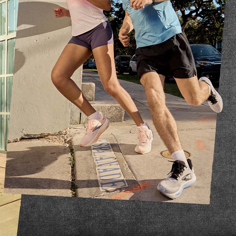 Two people running on a sidewalk wearing Brooks Ghost 18 running shoes with a building and parked cars in the background.