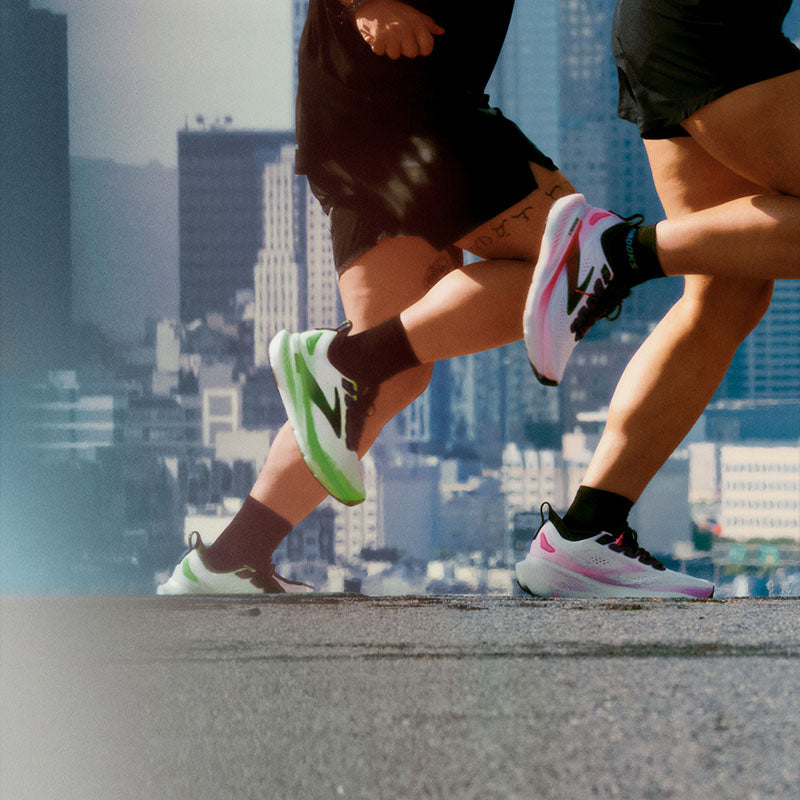 Two runners in white Brooks Glycerin 23 running shoes with pink and green accents against a city skyline.
