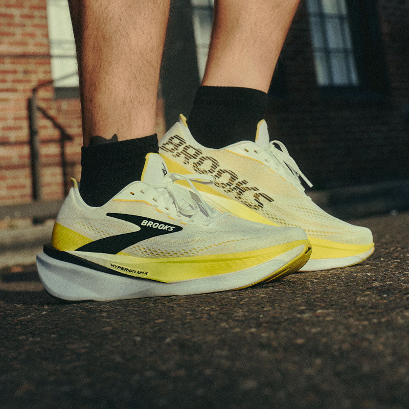 A runner wearing men's Brooks Hyperion Max 4 shoes on a paved surface with a brick building in the background