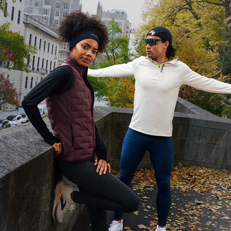 Two people wearing Brooks running clothing, headwear and shoes for fall weather, outdoors in a city with trees and buildings in the background