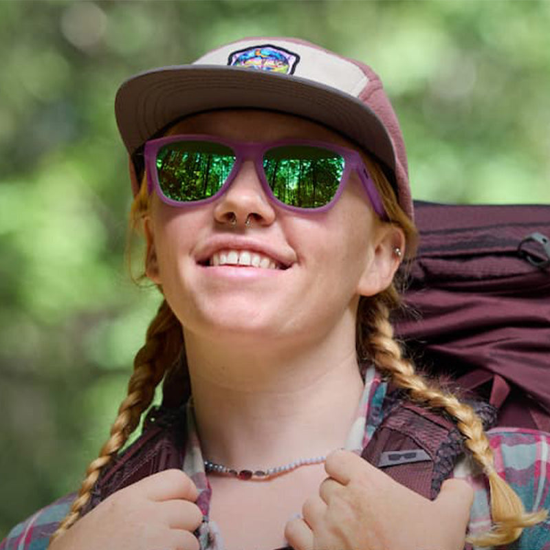 A person in a flannel shirt, trucker hat and purple goodr Sunglasses with green lenses wearing a large backpack in the woods with a blurred background