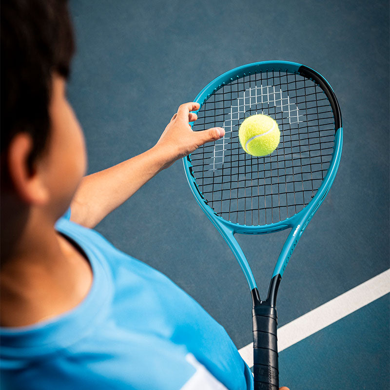 Over-the-shoulder view of a child holding a blue HEAD Boom junior tennis racquet on a blue court