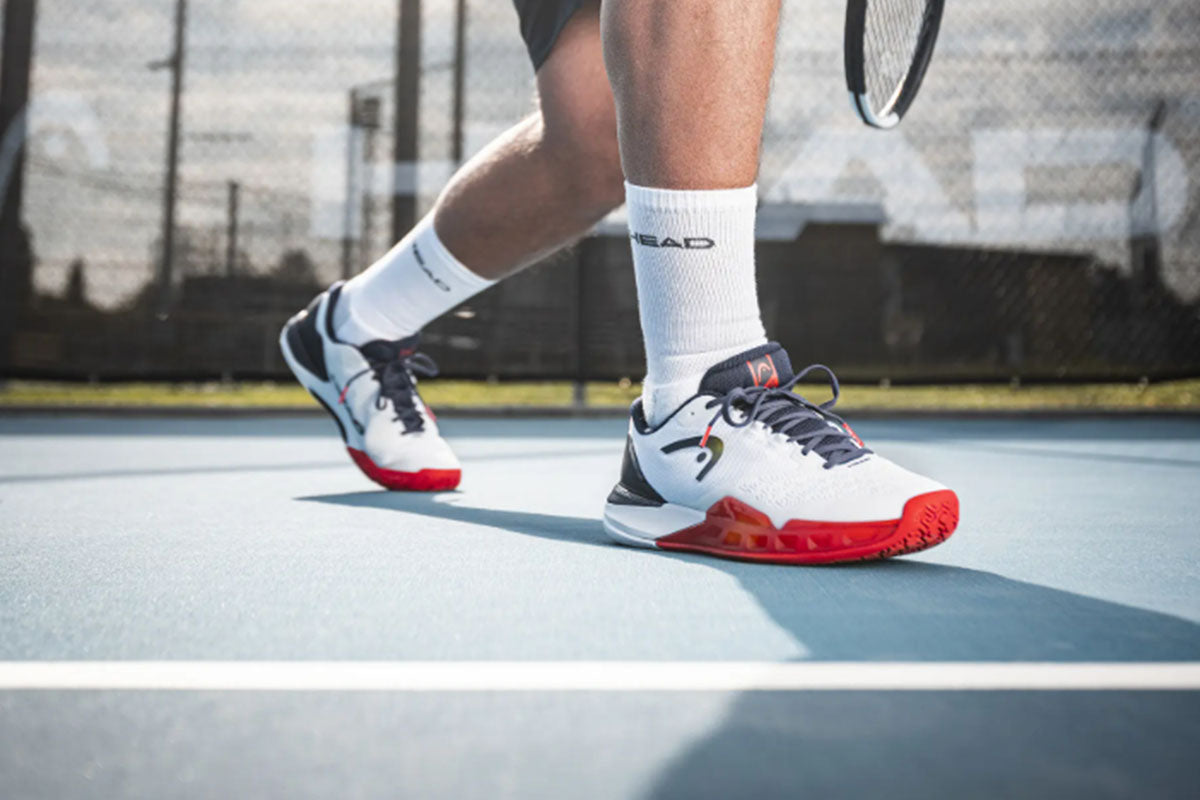 Tennis player wearing white HEAD Revolt Pro 5.0 tennis shoes with dark blue and red accents on a blue court with a blurred background