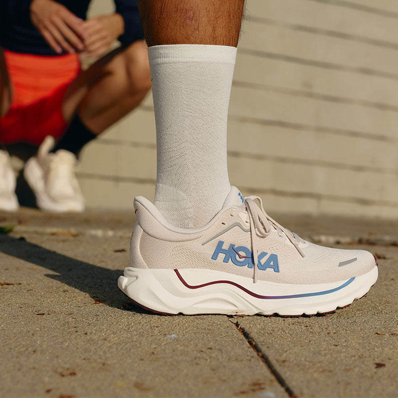 A close-up of a person wearing HOKA Arahi  9 running shoes and white crew socks on a concrete sidewalk. The shoes are off-white with blue and dark red accents. There is another person in the blurred background, leaning against an off-white brick wall.