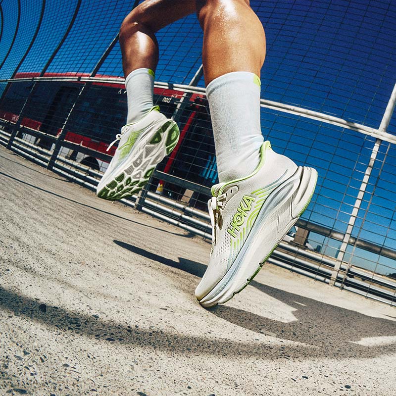 A runner wearing light colored HOKA Clifton 11 shoes with green and gray-blue accents with a tall wire fence and public transportation bus in the background