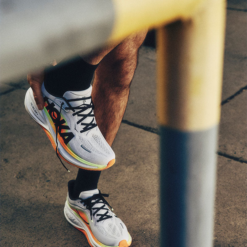 a runner putting on white HOKA Clifton Pro shoes with orange and yellow-green accents on a concrete sidewalk with a blurred structure in the foreground