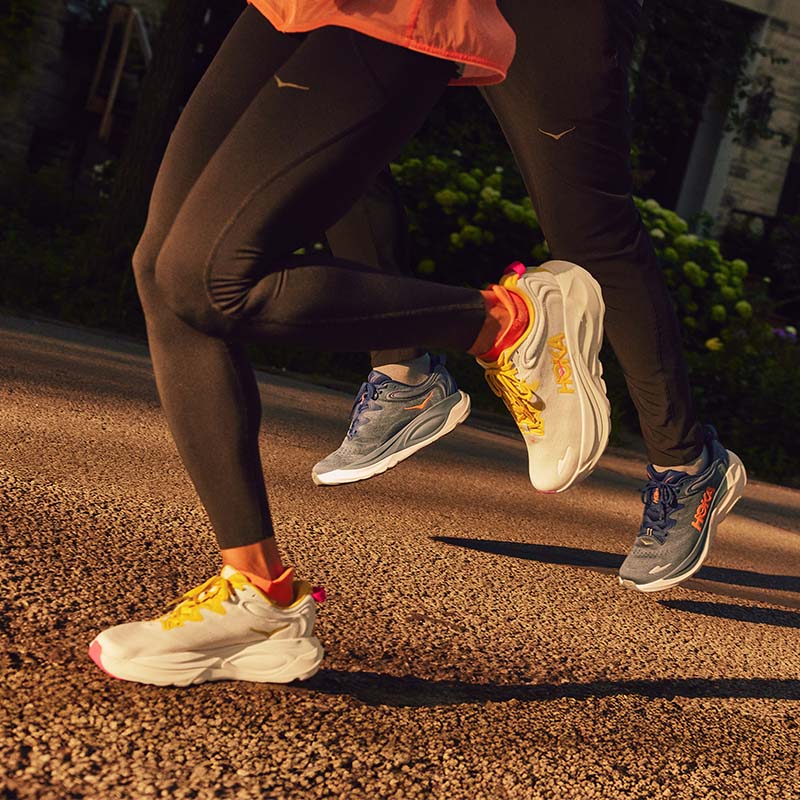 Two people running in HOKA Gaviota 6 running shoes on a road in front of a building with a lush flower garden