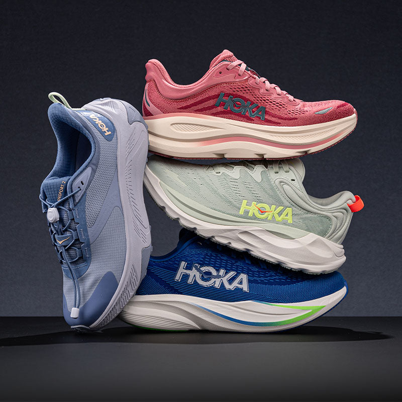 Four HOKA running shoes stacked and posed together on a dark studio background. The shoes show different colorways: light blue, dusty pink, pale mint green, and royal blue, each with contrasting lightly colored midsoles and bright accent colors.