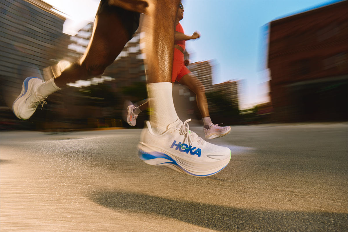 Two runners wearing men's and women's HOKA Skyward X 2 running shoes on a paved road against a motion-blurred cityscape background