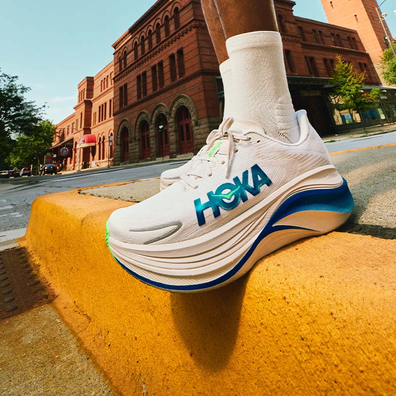 A person wearing white HOKA Skyward X 2 running shoes with blue and green accents on a yellow curb, city street background with red brick buildings