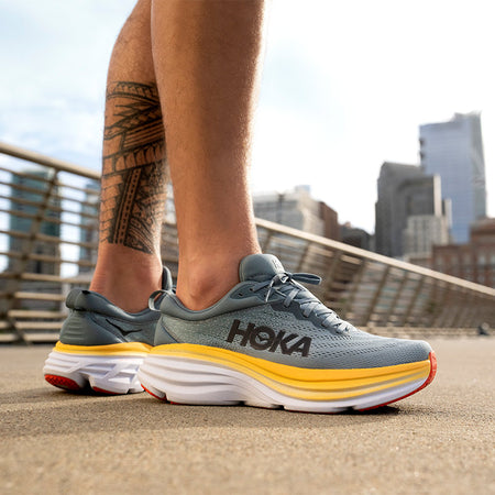 A person standing in men's HOKA Bondi 7 running shoes on a paved surface against a sunny cityscape.