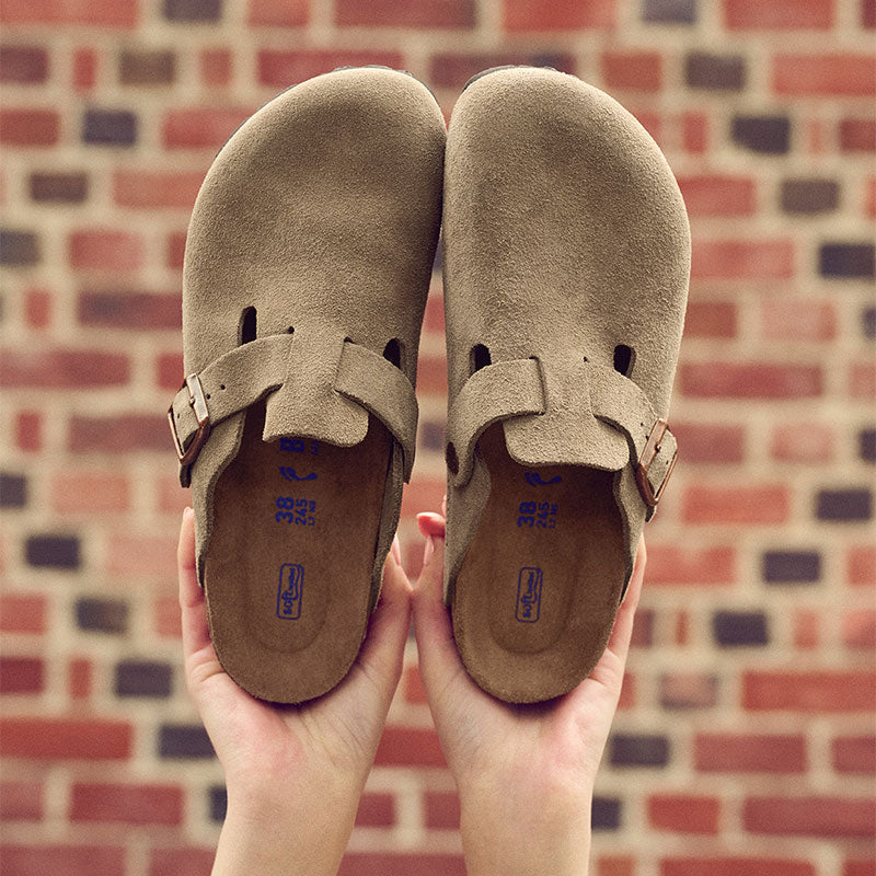 Closeup of two hands holding a pair of Taupe Birkenstock Boston Slides against a blurred, red brick wall background.
