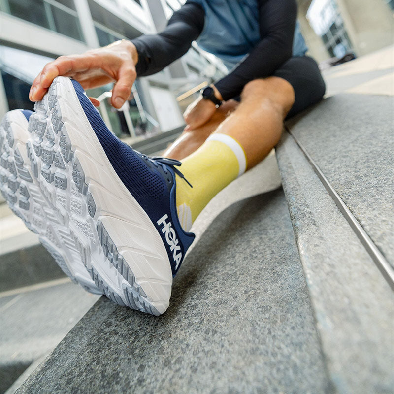 Lifestyle image: a person in navy blue and white HOKA Arahi 7 running shoes sitting and stretching by holding the toe of their right shoe with their left hand.