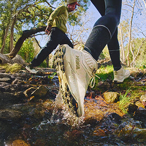 Two people hiking through a stream wearing HOKA Transport Hike GTX shoes.