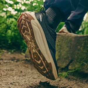 Person wearing a black and brown HOKA Transport 2 hiking shoe on a natural path with greenery.