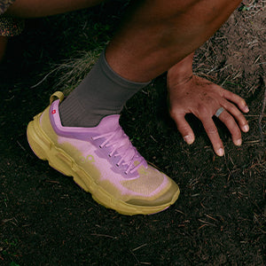 A person running a dirt trail in pink, purple and tan On Cloudsoma trail running shoes 