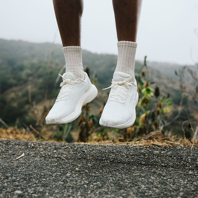 A person mid-jump wearing all-white hylo athletics running shoes and crew socks on a paved road with mountains in the background.