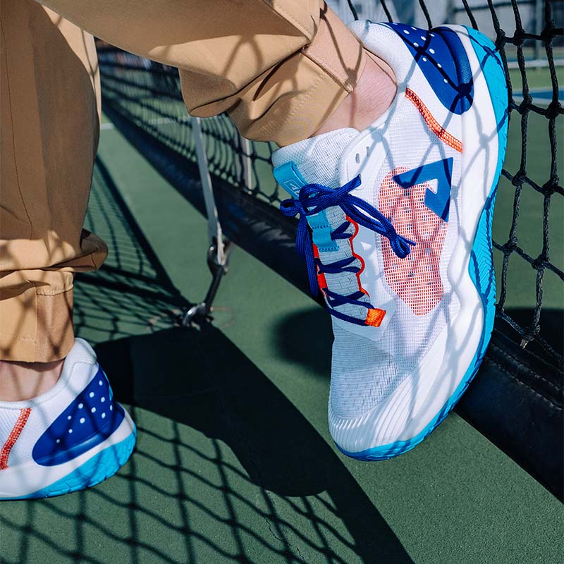 Close-up of a person wearing white, blue and orange JOOLA R4LLy Pickleball shoes next to a net on a green outdoor court surface.