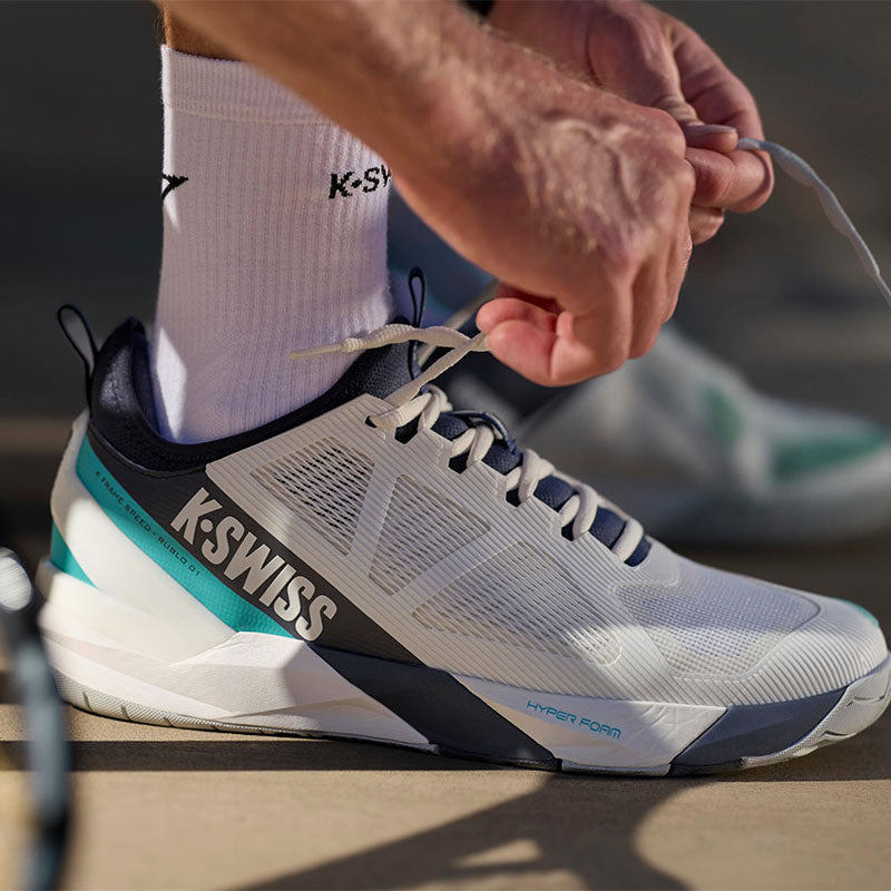 A close-up of a person tying the laces of a K-Swiss tennis shoe. The shoe is white with dark navy and teal accents, worn with white K-Swiss socks in an outdoor setting.