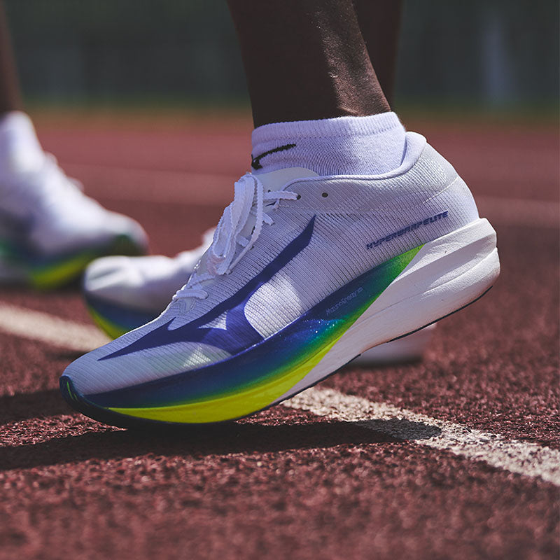 Closeup of a person wearing white Mizuno Hyperwarp Elite running shoes with blue and green accents on a red all-weather track with white lines