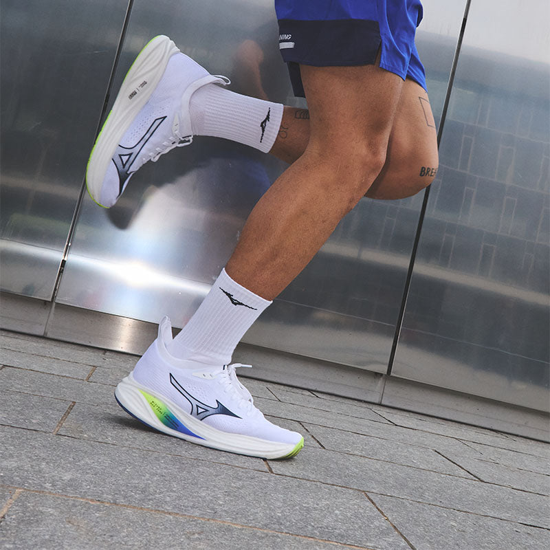 Person running in white Mizuno Neo Zen men's running shoes with colorful midssole accents, white socks, and blue athletic shorts against a reflective metallic wall.