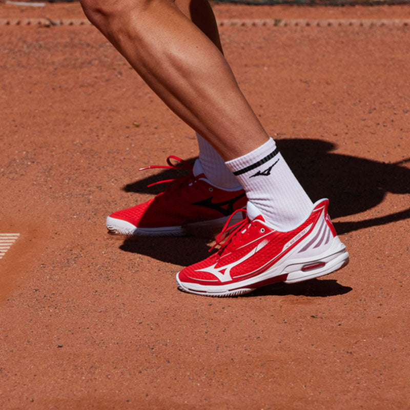 Tennis player wearing red Mizuno Wave Exceed Tour 7 AC men's tennis shoes with white accents on a clay court.