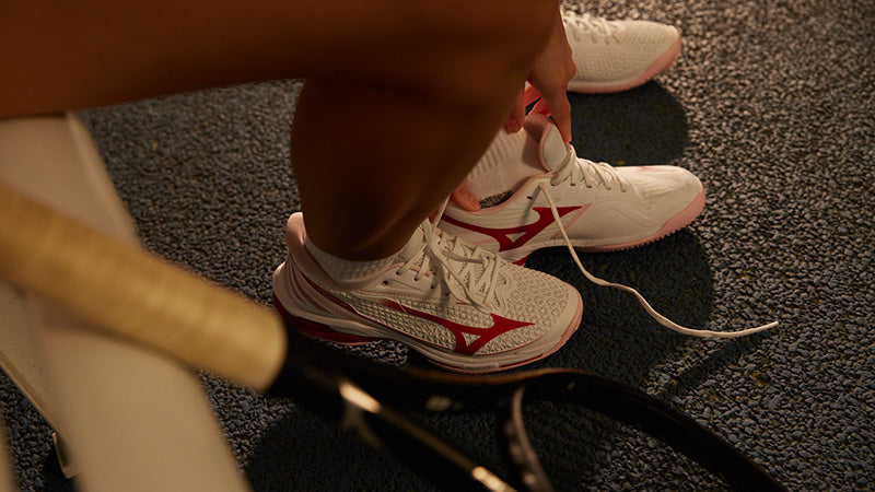 A person putting on a pair of white, red and pink Mizuno Wave Exceed Tour 7 women's tennis shoes