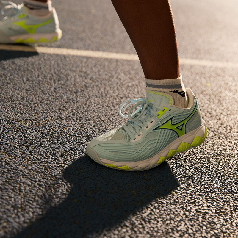 closeup of a person's feet wearing light blue and green Mizuno Wave Enforce Tour 2 AC women's tennis shoe on a dark gray court