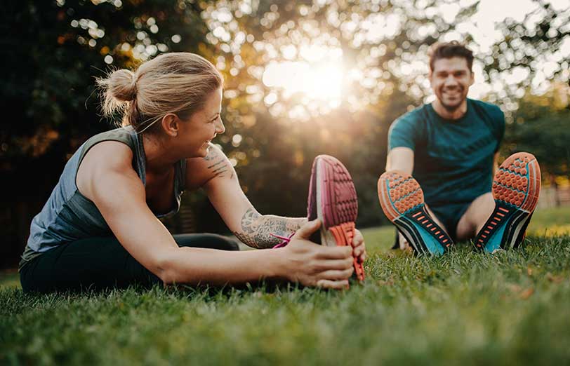 Woman and man seated on grass wearing athletic shoes and smiling at one another.