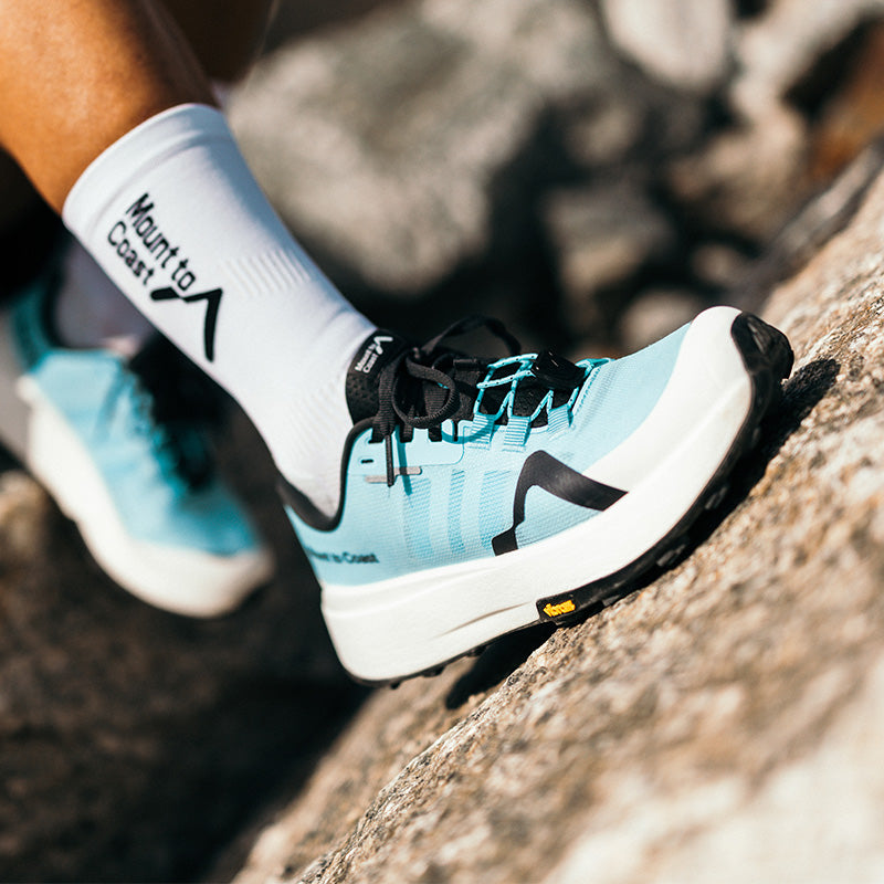 A close-up of a person wearing glacier blue Mount to Coast M1 trail running shoes on a rocky outdoor surface.