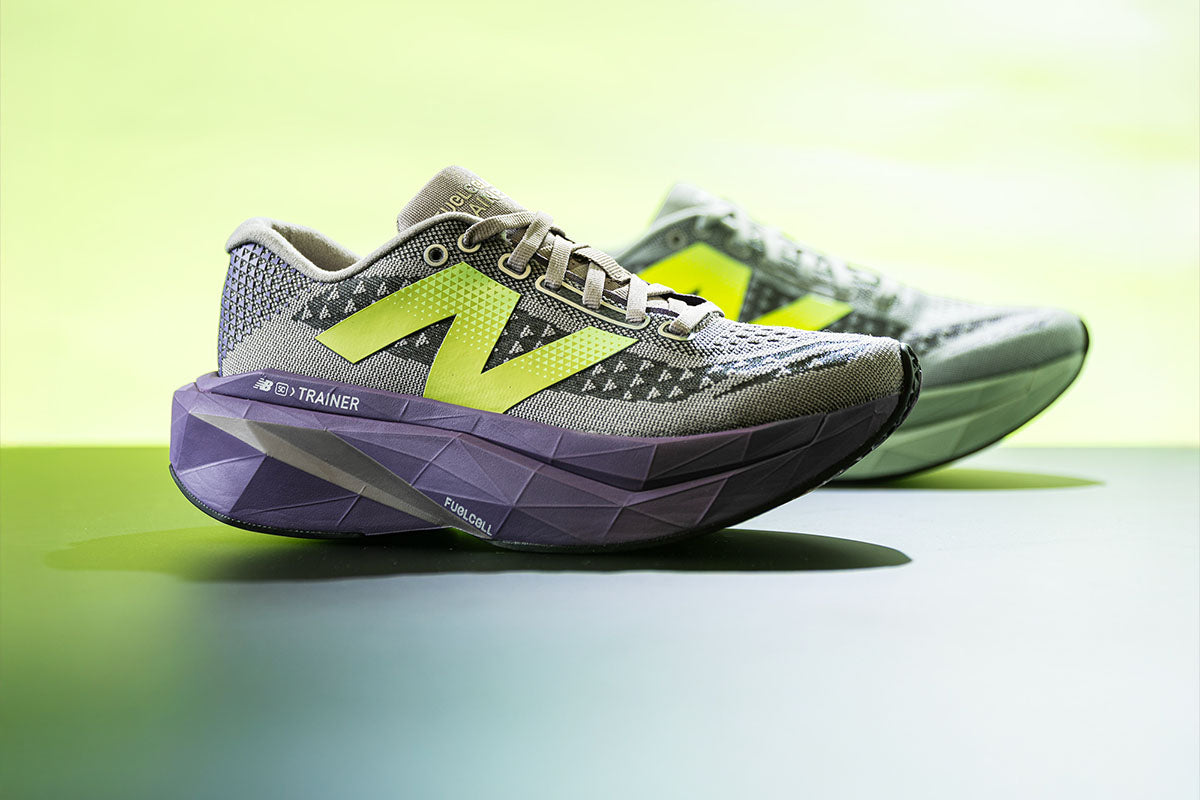 One gray and purple New Balance SuperComp Trainer v3 running shoe positioned in front of a blurred green shoe of the same model on a green background 