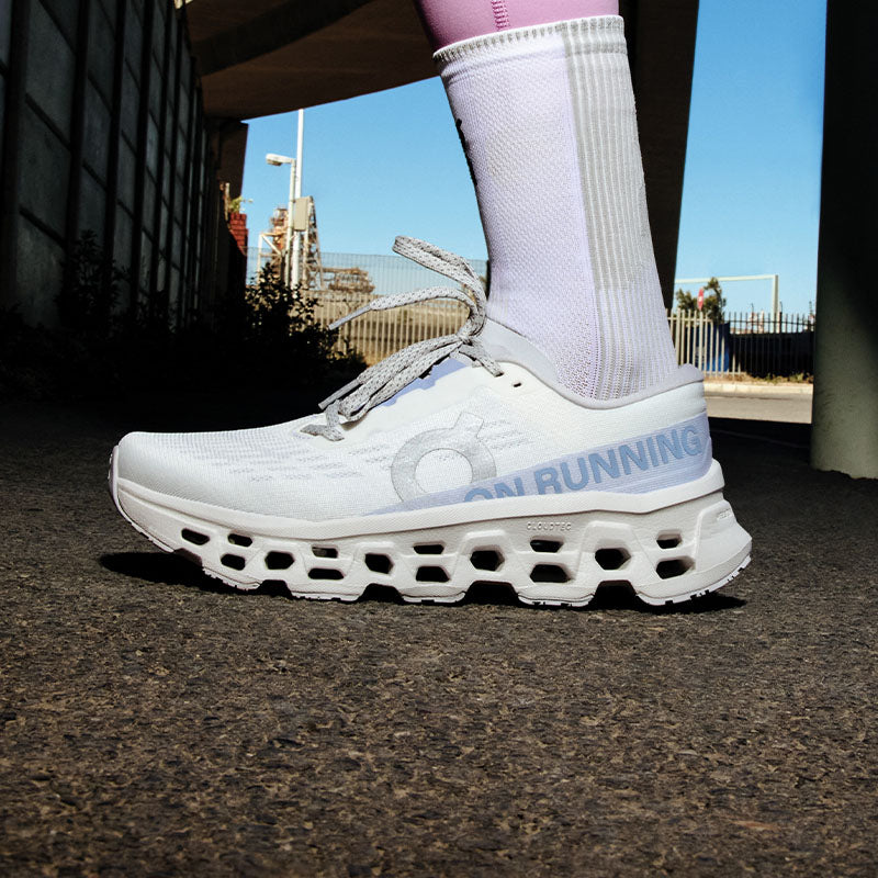 Closeup of a person wearing white and light blue On Cloudmonster 3 running shoes on urban pavement with a construction site in the background.